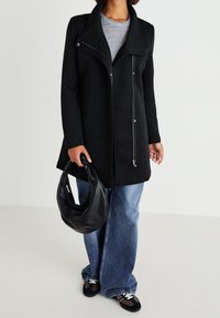 Zwarte ritssluitingjas met hoge kraag, gecombineerd met ruime lichtblauwe jeans en een slappe zwarte handtas. Heeft een gladde stof en een stijlvolle uitstraling.