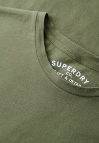 Camiseta de algodón verde oliva con cuello redondo, que presenta un logo blanco impreso en la etiqueta. Textura suave y diseño minimalista.