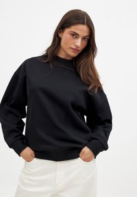 Sudadera negra de cuello redondo con mangas largas, puños y dobladillo acanalados. Tela suave con un ajuste relajado, combinada con pantalones de color claro.