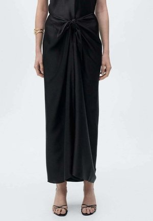 Vrouw draagt een zwarte lange rok met een knoopdetail aan de voorkant, gecombineerd met zwarte open teen sandalen met bandjes en een gouden armband aan haar linkerarm.