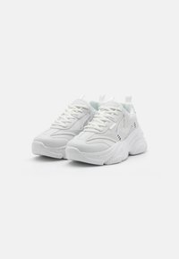 White athletic sneakers with a chunky sole, textured panels, flat laces, and subtle branding details on the sides and tongue.