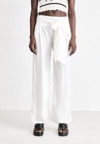Pantaloni larghi in lino bianco con dettaglio della vita legato, abbinati a sandali platform neri. Texture liscia, vestibilità comoda e design minimalista.
