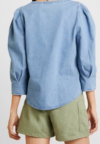 Top in denim azzurro chiaro con maniche puff a tre quarti, orlo curvo e dettagli sul colletto posteriore, abbinato a pantaloni corti verdi oliva.