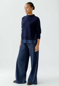 Marineblauer Strickpullover mit geripptem Halsabschuss, über ein Jeanshemd getragen. Kombiniert mit weitem Jeanshosen mit ausgefranstem Saum und dezenten Nähten.
