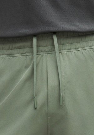 Nærbillede af olivengrønne bukser med elastik i taljen og matchende snørebånd samt en grå tekstureret skjorte stukket ind ovenover.