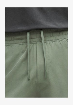 Nærbillede af olivengrønne bukser med elastik i taljen og matchende snørebånd samt en grå tekstureret skjorte stukket ind ovenover.