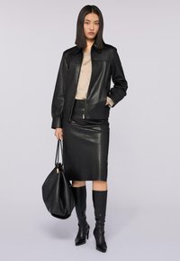 Giubbotto di pelle nera con zip frontale, abbinato a una gonna a tubino di pelle nera. La modella tiene una grande borsa nera a tracolla e indossa stivali neri fino al ginocchio.