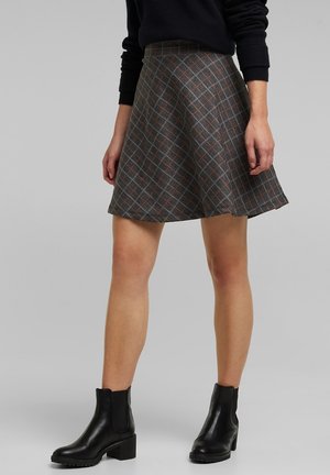 Kvinde iført et gråt ternet A-linje nederdel, sort sweater og sorte ankelstøvler, stående mod en ensfarvet baggrund.