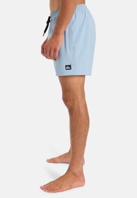 Light blue swim shorts made of textured fabric, featuring an elastic waistband and a black brand logo tag on the side.