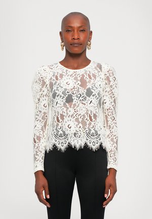 Woman with shaved head wears gold hoop earrings, a white floral lace long-sleeve top, and black fitted pants, standing against a plain background.