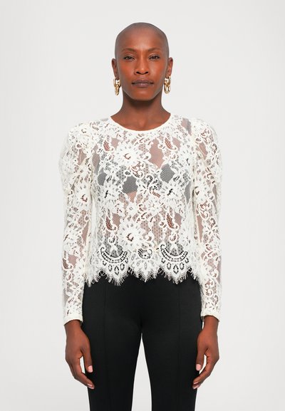 Une femme avec la tête rasée porte des boucles d'oreilles en or, un haut à manches longues en dentelle florale blanche et un pantalon noir ajusté, se tenant devant un fond uni.