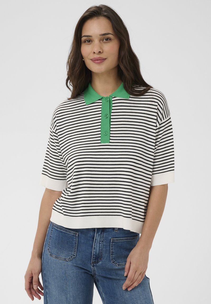 Jeune femme aux longs cheveux bruns portant un haut à manches courtes rayé blanc et noir avec un col vert et un jean bleu.