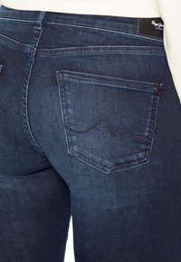 Primo piano di una tasca posteriore di jeans in denim blu scuro con un sottile design ricamato e un'etichetta nera di Pepe Jeans London sulla vita.