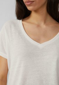 T-shirt blanc à manches courtes avec un col en V. Le tissu semble léger et légèrement texturé, offrant une coupe décontractée.
