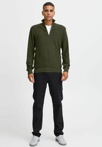 Dunkelgrüner, strukturierten Pullover mit einem Viertelreißverschluss-Hals, kombiniert mit schwarzen Hosen und grauen Sneakers. Der Pullover hat lange Ärmel und ein körperbetontes Design.