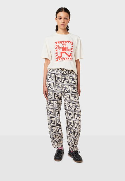 White short-sleeve t-shirt with red graphic design, paired with patterned cream pants featuring blue geometric shapes. Black shoes with pink accents.