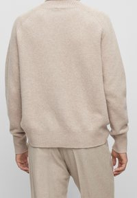 Beige stickad tröja med hög krage och ribbade ärmslut, med en avslappnad passform och textil med struktur, visad bakifrån.