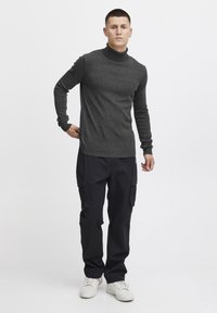 Grauer Rollkragenpullover kombiniert mit schwarzen Cargo-Hosen, die Taschen haben. Das Model trägt weiße Sneakers und steht vor einem schlichten Hintergrund.