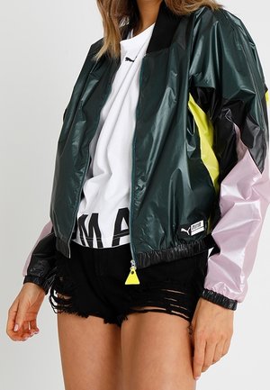 Mujer con una chaqueta Puma de color verde oscuro, negro, amarillo y rosa sobre una camiseta blanca de Puma y pantalones cortos negros rasgados.