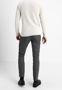 Světle béžový top s dlouhým rukávem a kulatým výstřihem s texturovaným vzorem, kombinovaný s tmavě šedými dobře padnoucími kalhotami na míru a černou obuví.