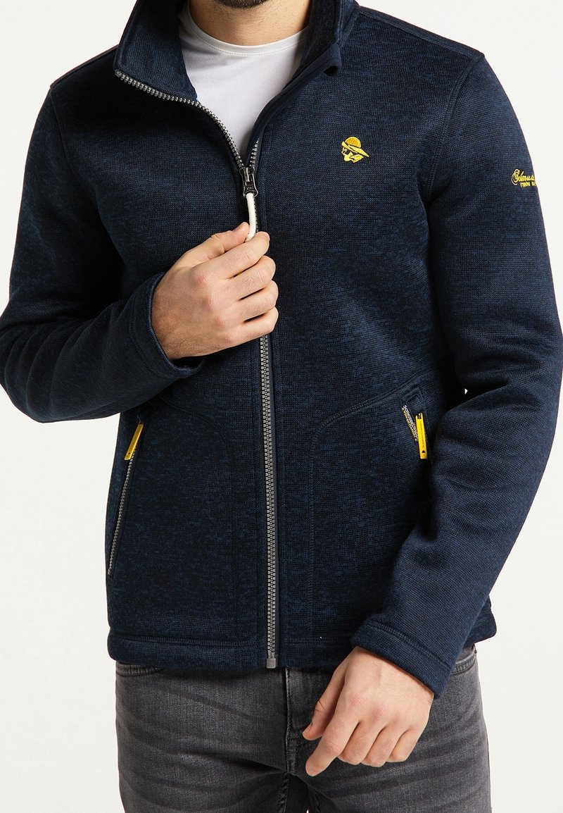 Homme portant une veste zippée bleu marine avec un logo jaune sur la poitrine et la manche, tenant la fermeture éclair près du col, vêtu d'un jean gris.