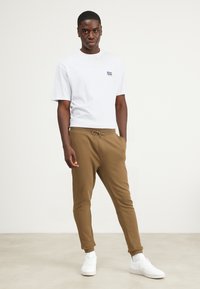 Witte T-shirt met korte mouwen en marineblauw logo, gecombineerd met bruine joggingbroek met elastische tailleband en trekkoord, afgemaakt met witte sneakers.