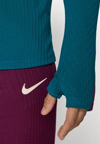 Teal geribbelde lange mouwen top met duimgatdetail, gecombineerd met kastanjebruine geribbelde broek met een crèmekleurig Nike-logo. Rekbare stof met een gestructureerde afwerking.