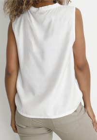 Bílý top bez rukávů s nařaseným výstřihem, vyrobený z lehkého materiálu. Nosen s světle béžovými kalhotami, které ukazují hladkou strukturu a čisté linie.