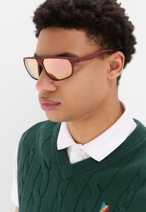Giovane uomo che indossa occhiali da sole specchiati con montatura color granata, camicia bianca con colletto e gilet di maglia verde scuro.