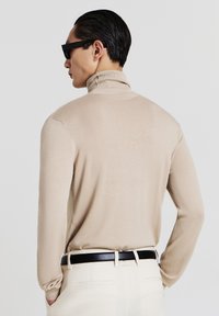 Beige turtleneck sweater with long sleeves, ribbed collar, and a smooth texture. Worn with cream-colored pants and black belt.