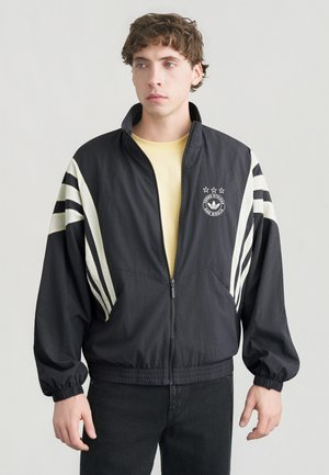 Veste coupe-vent noire avec des bandes blanches sur les manches, poignets et ourlet élastiques, fermeture éclair avant et logo sur la poitrine.