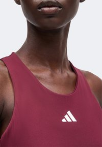 Gros plan d'une personne portant un haut sans manches bordeaux avec un logo blanc Adidas sur la poitrine, montrant les épaules et le bas du visage.