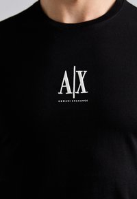 Black cotton t-shirt featuring a white embroidered logo "A/X" with "ARMANI EXCHANGE" text below. Crew neckline, smooth texture, fitted design.