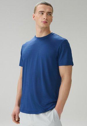 Young man with short hair wearing a fitted blue short-sleeve shirt and white pants, looking sideways with one hand in pocket.