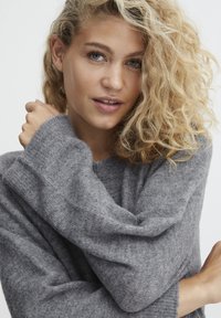 Pulover gri din tricot cu mâneci largi, textură moale și croială lejeră. Modelul are părul creț și își încrucișează ușor brațele.