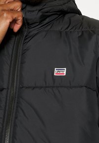 Svart vadderad jacka med hög krage, utrustad med en dragkedja framtill och en liten rektangulär Levi's-logotyp på bröstet. Mjukt, lätt material.