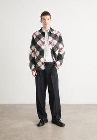 Plaid jacket in black, white, and burgundy with two chest pockets, worn over a white shirt. Black trousers and shoes complete the outfit.
