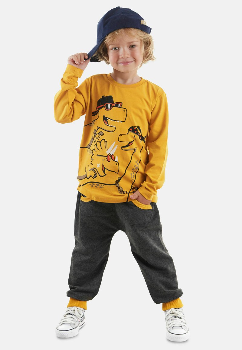 Geel langok mouw shirt met dinosauruss grafieken; grijze joggingsbroek met gele boorden; witte sneakers; marineblauwe pet achterstevoren gedragen.