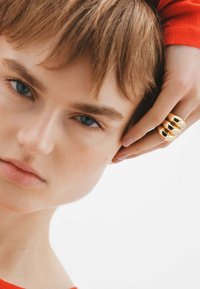 Anillo de metal dorado con tres secciones redondeadas, acabado pulido. La mano del modelo descansa contra una oreja, con un suéter rojo visible en el fondo.