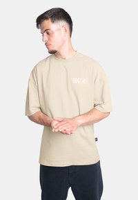 Beige oversized tričko vyrobené z bavlny, s krátkými rukávy a na levém prsu je bíle vytištěno slovo "CRYSTAL".