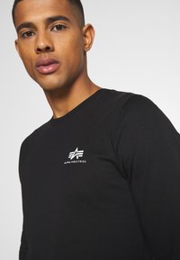 Schwarzes Langarm-T-Shirt aus Baumwolle mit taillierter Form, ausgestattet mit einem weißen Logo auf der linken Brustseite. Glatte Textur und einfaches Design.