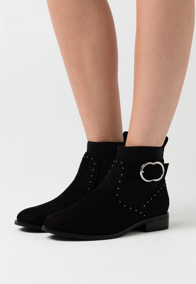 Bottines en daim noir avec panneaux latéraux élastiques, détails en métal argenté et clous argentés le long des côtés. Talon plat et bas.