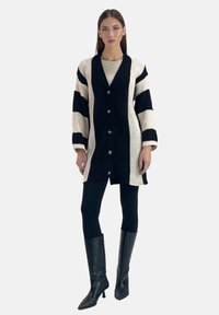 Svart och krämrandig cardigan med en texturerad stickad design, frontknappar och trekvartslånga ärmar, parat med svarta leggings och knähöga stövlar.