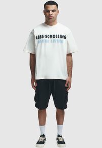 T-shirt oversize blanche avec du texte noir et bleu portant la mention "MOINS DE DÉFILEMENT, PLUS DE VIE", associée à un short cargo noir et des baskets noires.