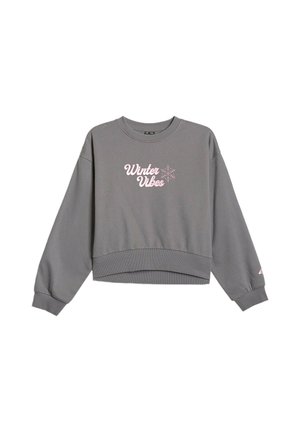 Dziewczyna z długimi brązowymi włosami ubrana w szarą bluzę i spodnie dresowe z różowym napisem „"Winter Vibes"” oraz czarno-białe sneakersy.