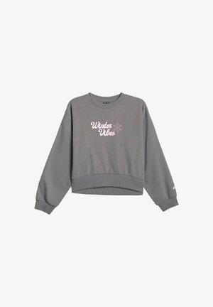 Dziewczyna z długimi brązowymi włosami ubrana w szarą bluzę i spodnie dresowe z różowym napisem „"Winter Vibes"” oraz czarno-białe sneakersy.