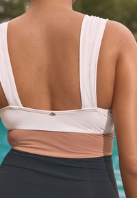 Vista posteriore di una persona che indossa un costume da bagno a blocchi di colore con spalline bianche, parte centrale beige e parte inferiore scura vicino a una piscina.