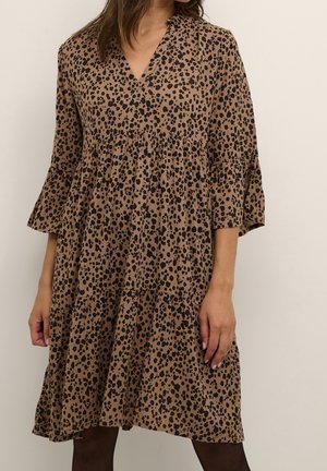 Frau trägt ein braunes, knielanges Kleid mit schwarzen unregelmäßigen Flecken und 3/4-Ärmeln, steht vor einem schlichten hellen Hintergrund.