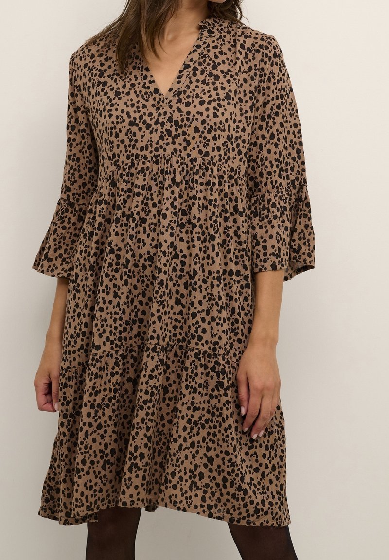 Femme portant une robe marron mi-longue avec des taches noires irrégulières et des manches trois-quarts, se tenant devant un fond clair uni.