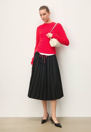 Femeie purtând un pulover roșu, fustă midi plisată neagră cu snur, pantofi negri cu toc și ținând o geantă mică albă de umăr.
